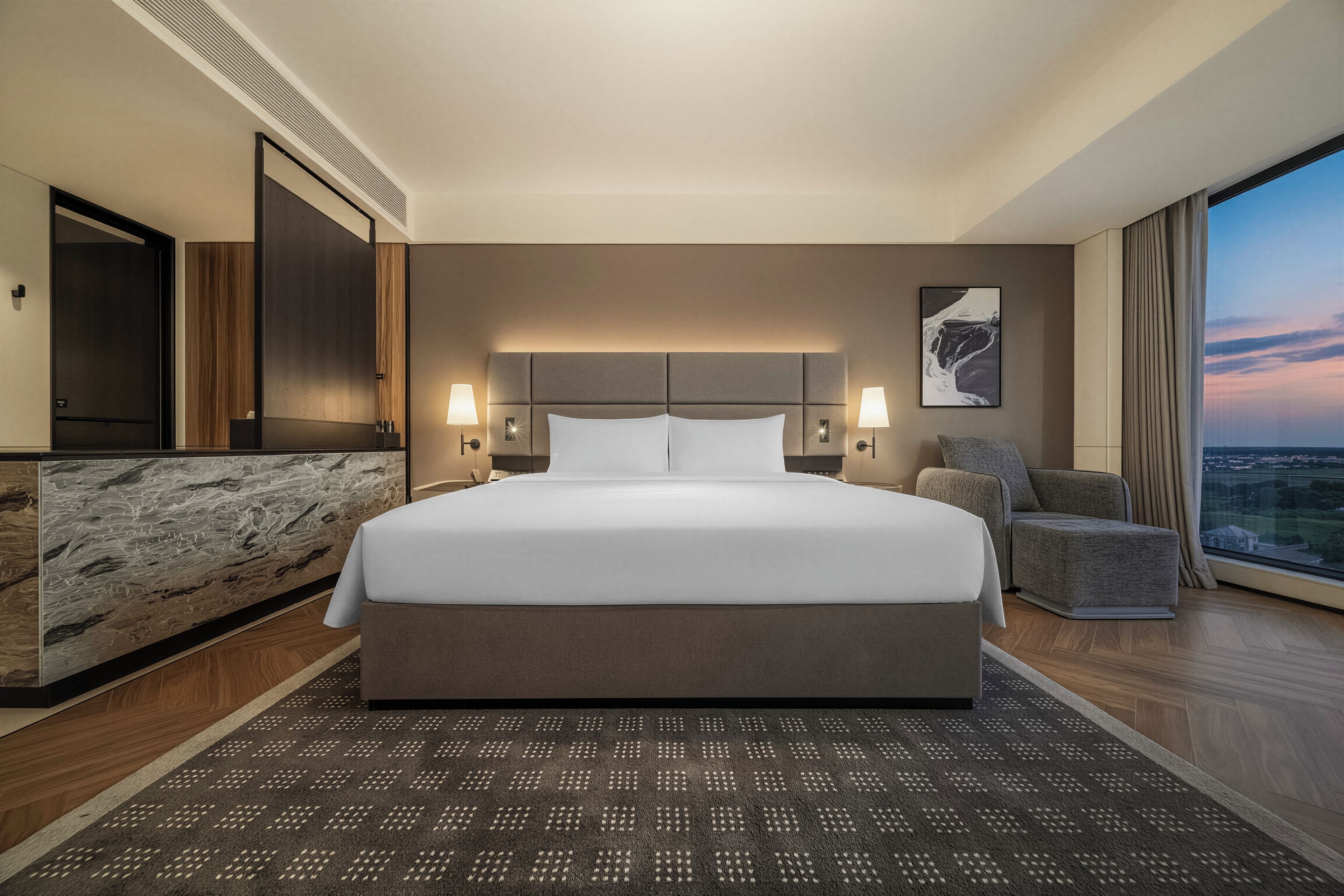 A hotel room with a large bed, a sofa, a view of the city, and a large window.