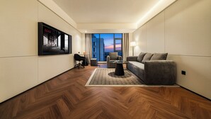 A modern hotel room with a large window, a sofa, a small table, and a TV mounted on the wall.