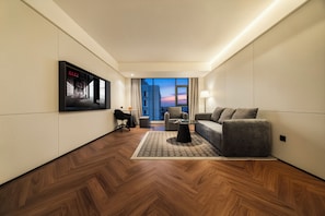 A modern hotel room with a large window, a sofa, a small table, and a TV mounted on the wall.