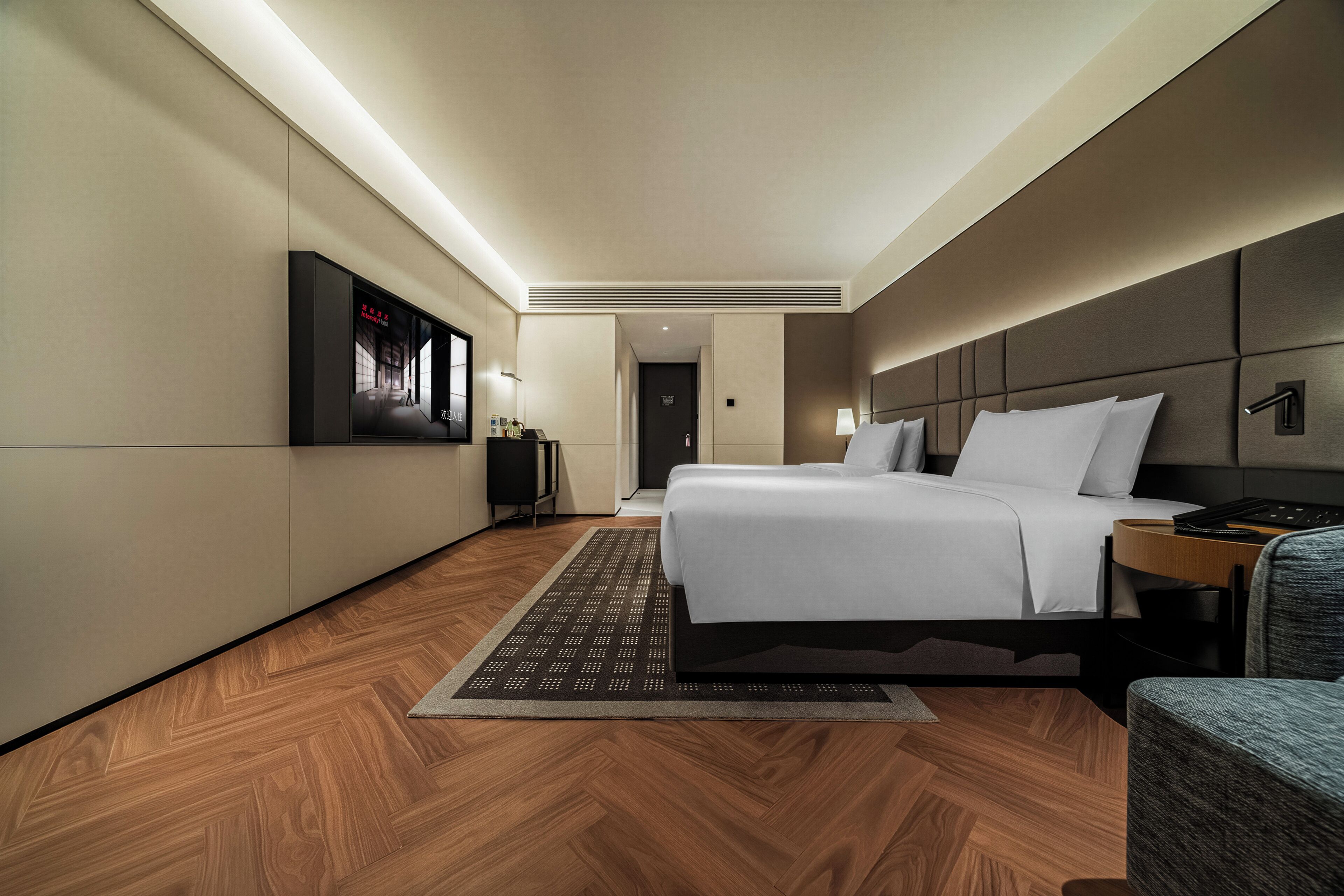 A modern hotel room with a large bed, a flat-screen TV mounted on the wall, a bedside table with a lamp, and a wooden floor.