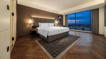 A modern hotel room with a large bed, a view of the cityscape through floor-to-ceiling windows, and a wooden floor.