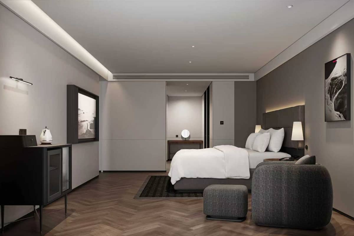 Ein modernes Hotelzimmer mit Bett, Nachttisch, Lampe und einem grauen Sessel.