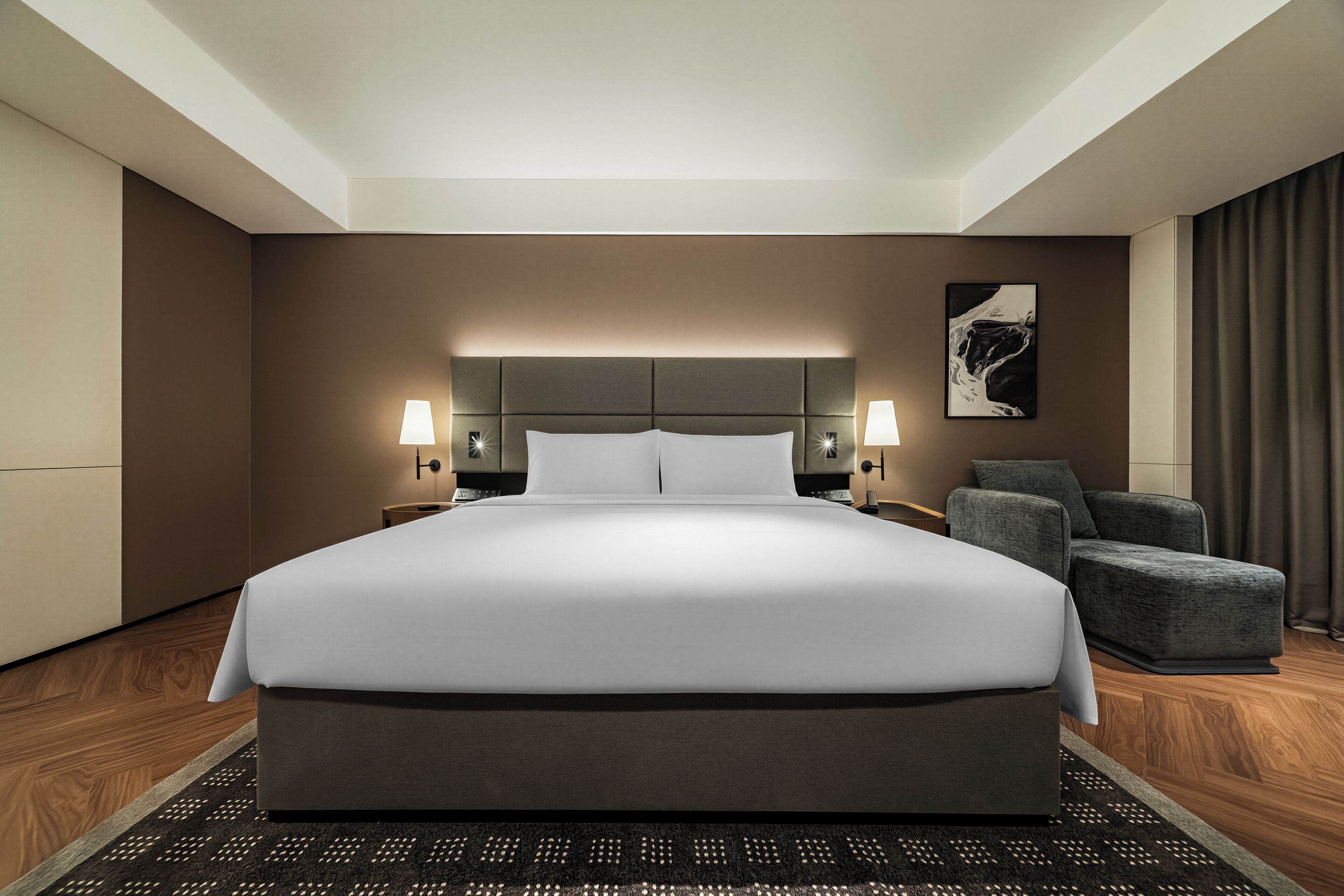 A modern hotel room with a large bed, two bedside lamps, a grey armchair, and a framed artwork on the wall.