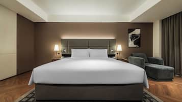 A modern hotel room with a large bed, two bedside lamps, a grey armchair, and a framed artwork on the wall.
