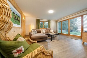Ein Wohnzimmer mit einer Couch, einem Couchtisch und einem Korbstuhl. An der Wand hängt ein Gemälde, und ein Fenster ist mit Vorhängen drapiert.