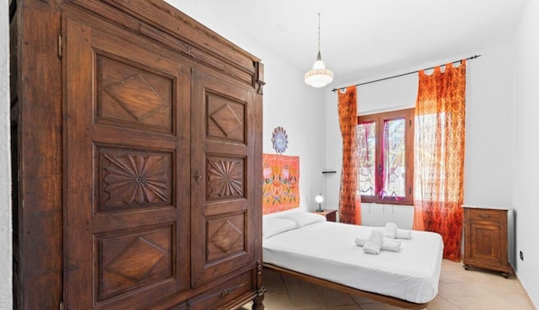 Un dormitorio con un armario grande de madera, una cama con sábanas blancas, una mesita de noche con una lámpara y cortinas anaranjadas.