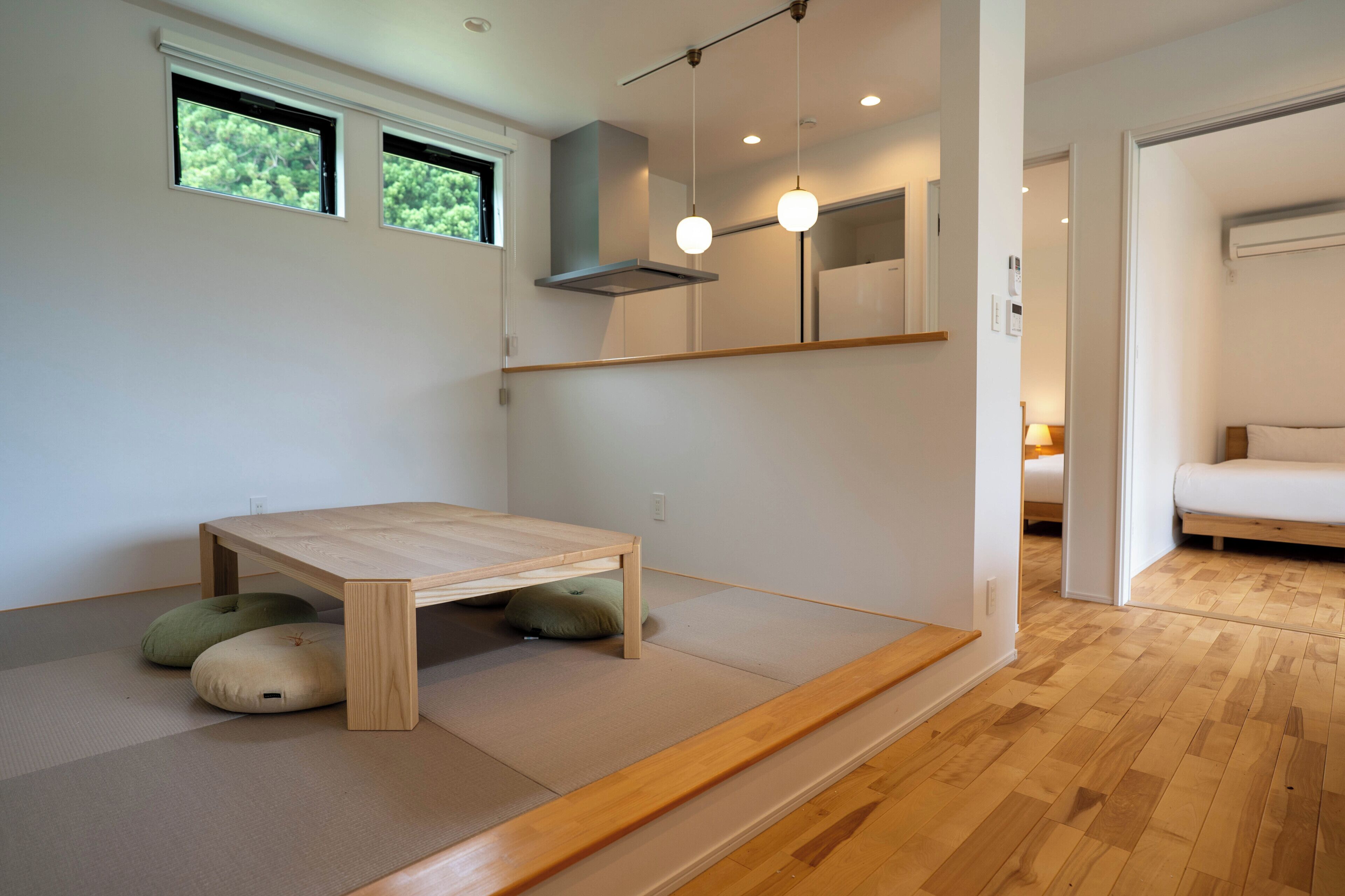 A minimalist room with a wooden table, two windows, a ceiling light, and a bed in the adjacent room.