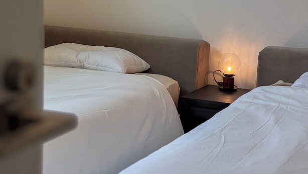A hotel room with two beds, a nightstand with a lamp, and a door partially open.