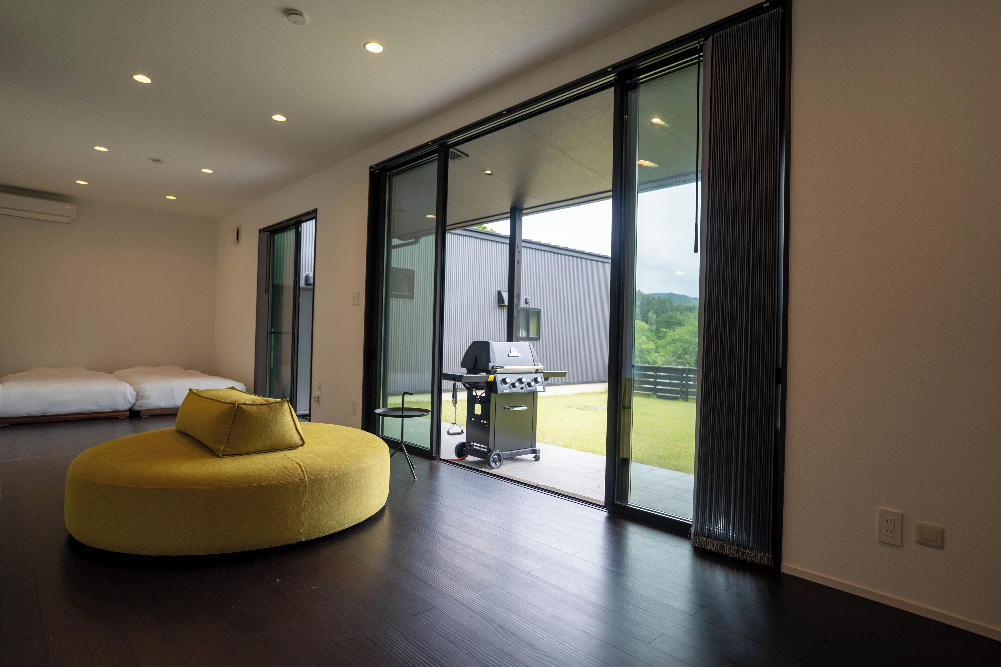 Una habitación moderna con un amplio ventanal corredera que da a un balcón, un otomano amarillo redondo y una pequeña mesa con una parrilla.