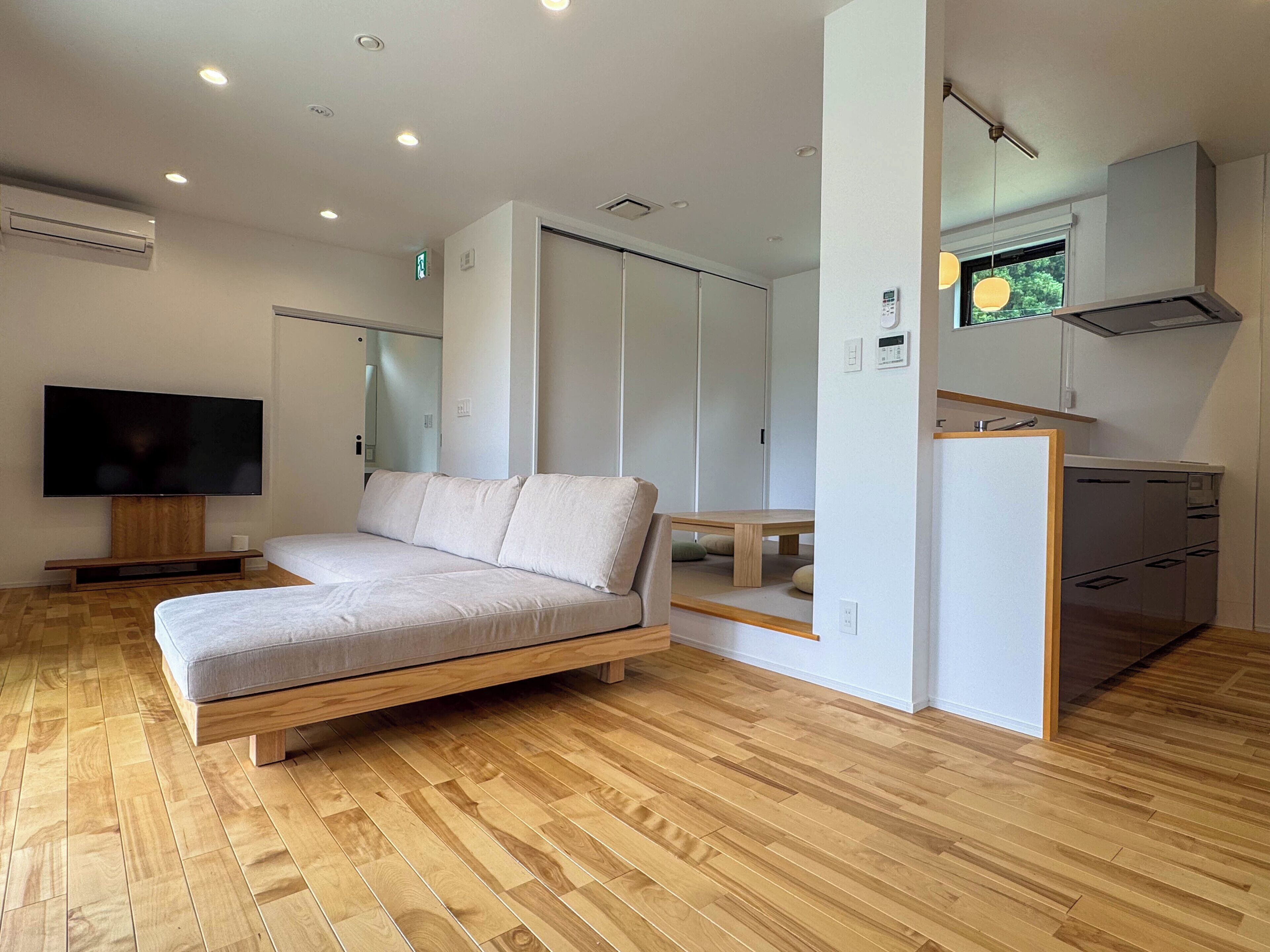 A modern hotel room with a wooden floor, a sofa, a television, a kitchenette, and a sliding door closet.