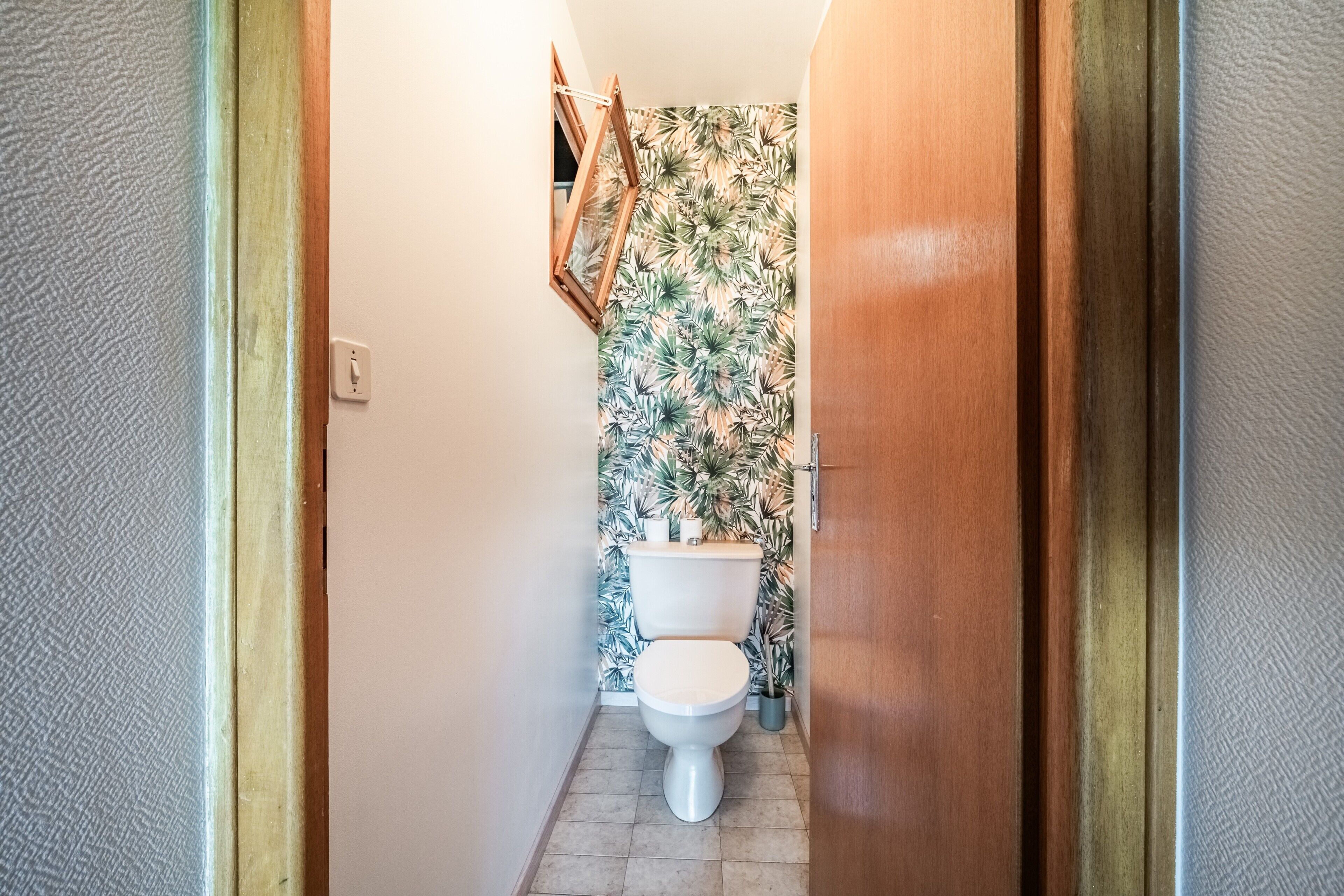 Ein schmaler Flur, der zu einem Badezimmer mit Toilette, Toilettenpapierrolle und WC-Bürste führt.