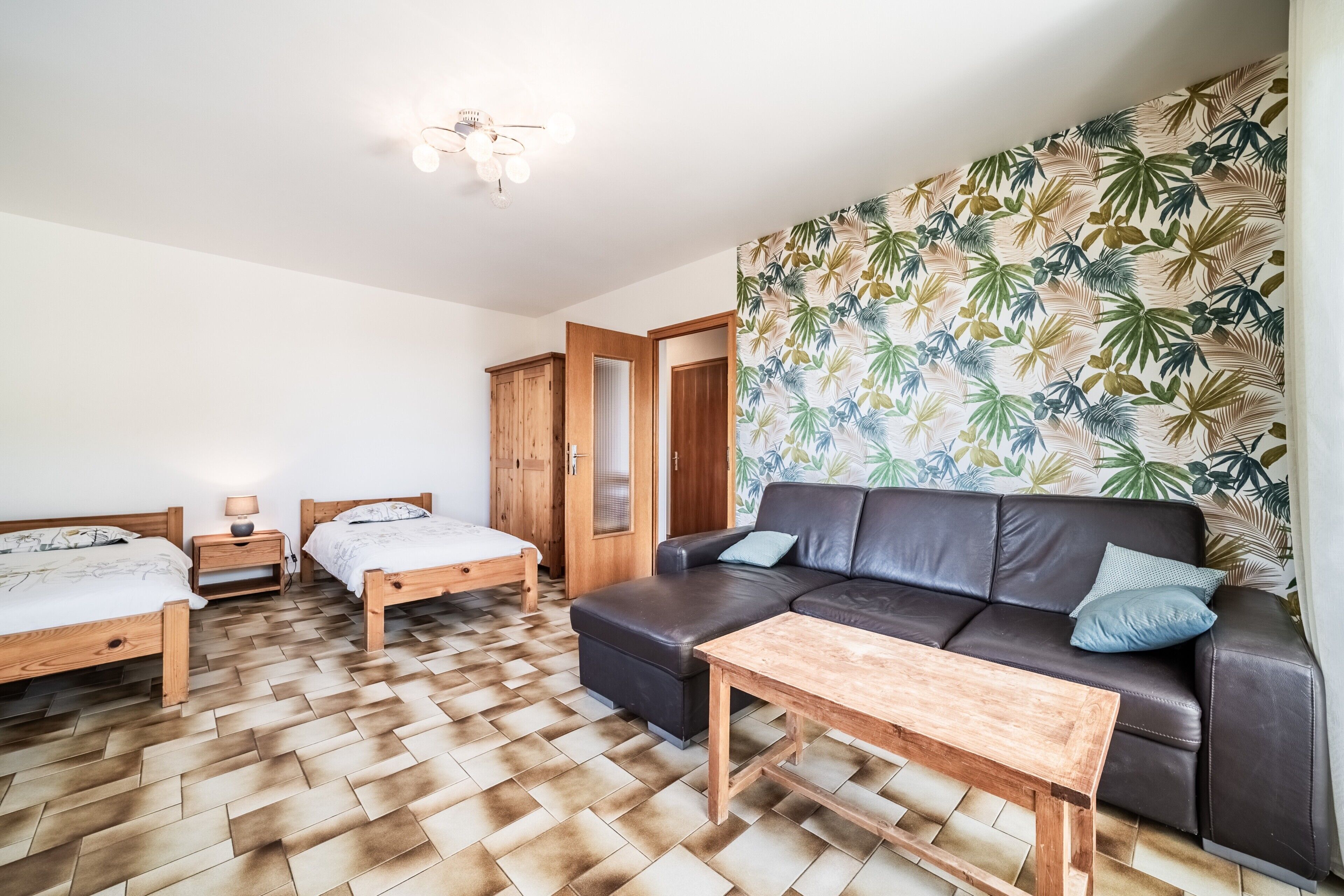 Une pièce dotée d’un parquet en chevrons, de deux lits en bois, d’un canapé en cuir, d’une table basse en bois et d’un mur orné d’un motif de feuilles tropicales.