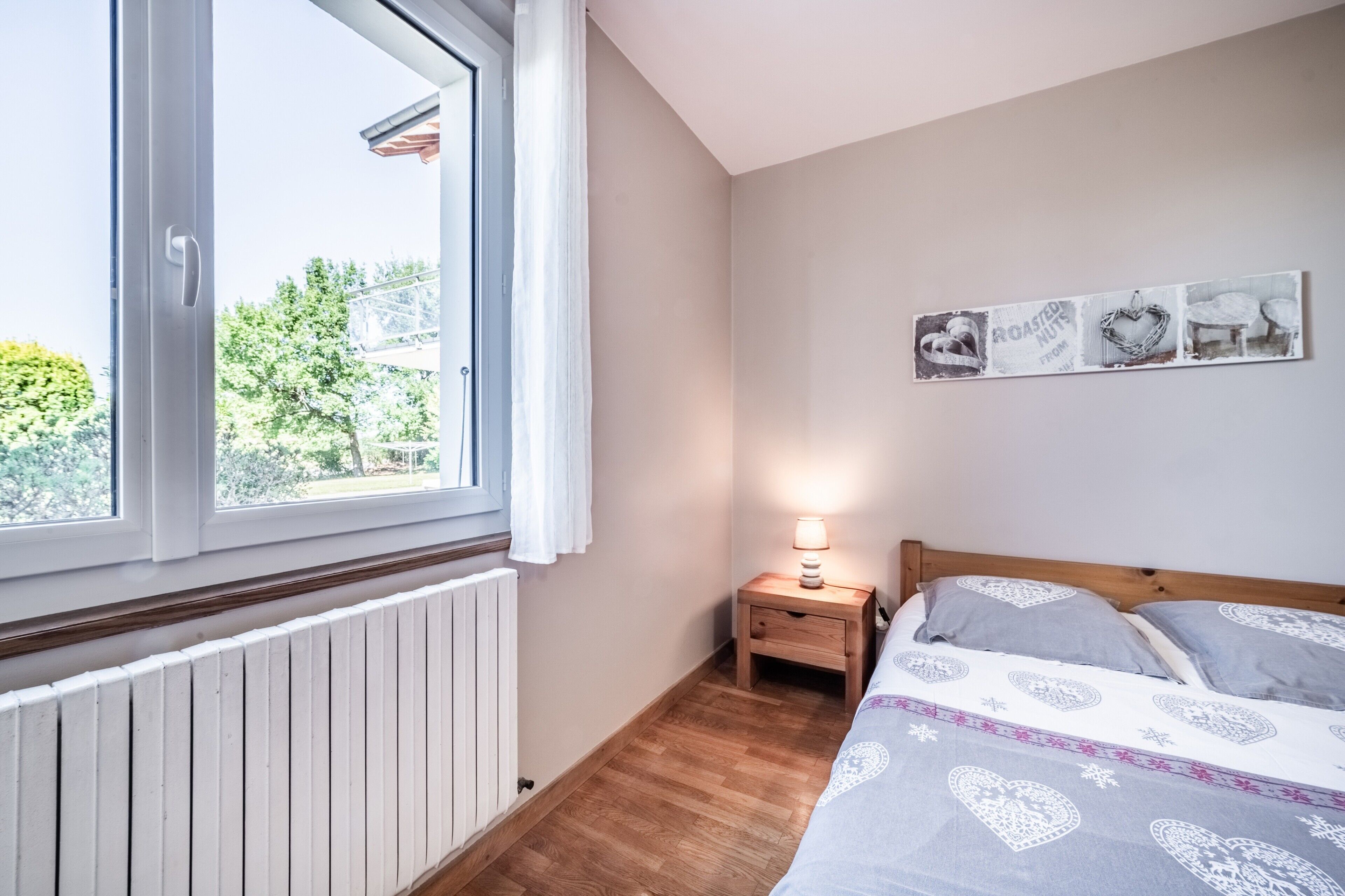 Et soveværelse med et stort vindue, et sengebord med en lampe og en seng med mønstret sengetøj.