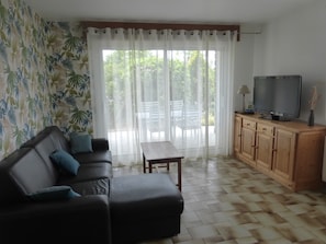 Ein Wohnzimmer mit einer Couch, einem Fernseher auf einer Holzkommode und Blick durch ein Fenster mit transparenten Vorhängen.