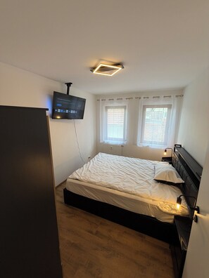 Ein Schlafzimmer mit Bett, einem an der Wand befestigten Fernseher, einem Fenster mit Vorhängen und einem Heizkörper unter dem Fenster.