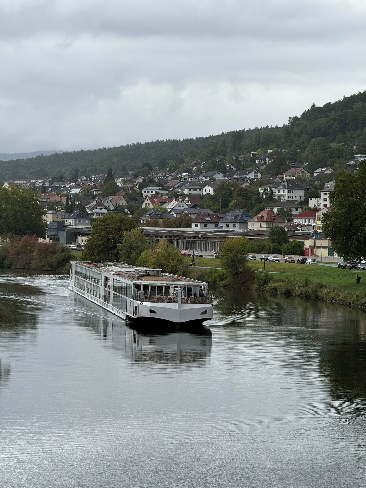 Un barco de paseo fluvial con cubierta plana y zona de estar, navegando por un río tranquilo con una zona residencial al fondo.