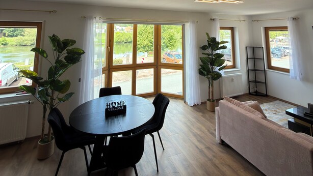 Um quarto espaçoso com uma mesa de jantar, cadeiras, um sofá e grandes janelas com vista para um lago e carros estacionados.