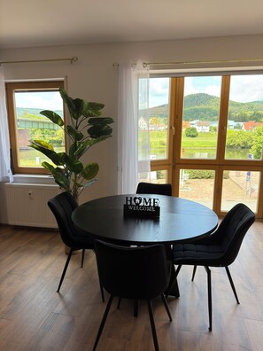 Ein Zimmer mit einem runden Tisch, vier Stühlen, einem Schild mit der Aufschrift 'HOME WELCOME', einer Topfpflanze und großen Fenstern mit Blick auf Grünflächen und Gebäude.
