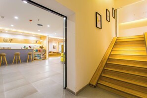A hotel lobby with a wooden staircase, a reception desk, and a seating area with stools.