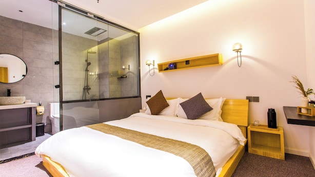Moderni hotellihuone, jossa on suuri sänky, lasinen suihkuseinä, puinen yöpöytä, pieni hylly ja seinään kiinnitetty valaisin.