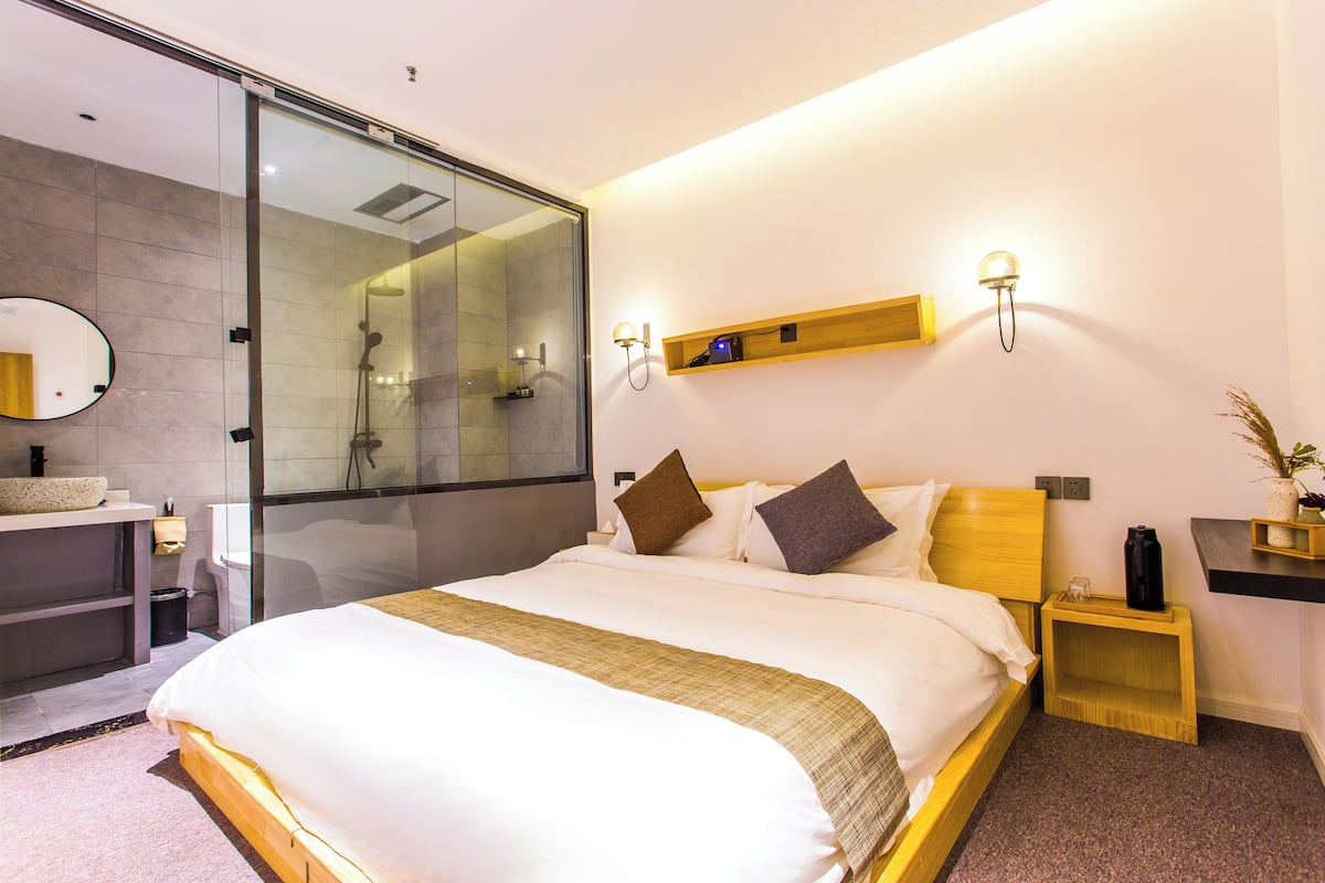 Una habitación de hotel moderna con una cama grande, un box de ducha de vidrio, una mesita de noche de madera, un estante pequeño y una lámpara fijada a la pared.
