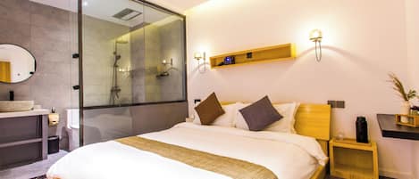 Una habitación de hotel moderna con una cama grande, un box de ducha de vidrio, una mesita de noche de madera, un estante pequeño y una lámpara fijada a la pared.