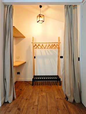 A room with a wooden floor, a hanging light fixture, a wooden clothes rack, and a shelf. - The Sun's Attic (Sestu)
