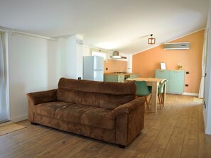 A living room with a brown sofa, a dining area with a table and chairs, and a kitchen with a refrigerator and stove. - The Sun's Attic (Sestu)