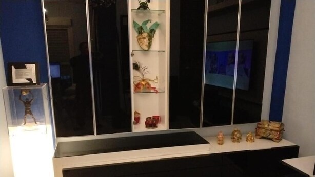 Ett modernt rum med ett svartvitt konsolbord, ett vitrinskåp i glas med dekorativa föremål och en tv på väggen.