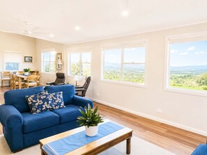 A spacious living room with a blue sofa, a dining area with a table and chairs, and large windows offering a view of the countryside. - Star of the Sea Milton Mollymook (Milton)
