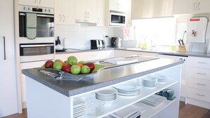 Una cocina moderna con armarios blancos, un horno de acero inoxidable, un microondas y un fregadero. Hay manzanas en la encimera y un escurreplatos con platos.
