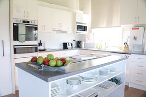 Eine moderne Küche mit weißen Schränken, einem Edelstahlbackofen, einer Mikrowelle und einem Spülbecken. Auf der Arbeitsfläche liegen Äpfel und ein Geschirrgestell mit Tellern.