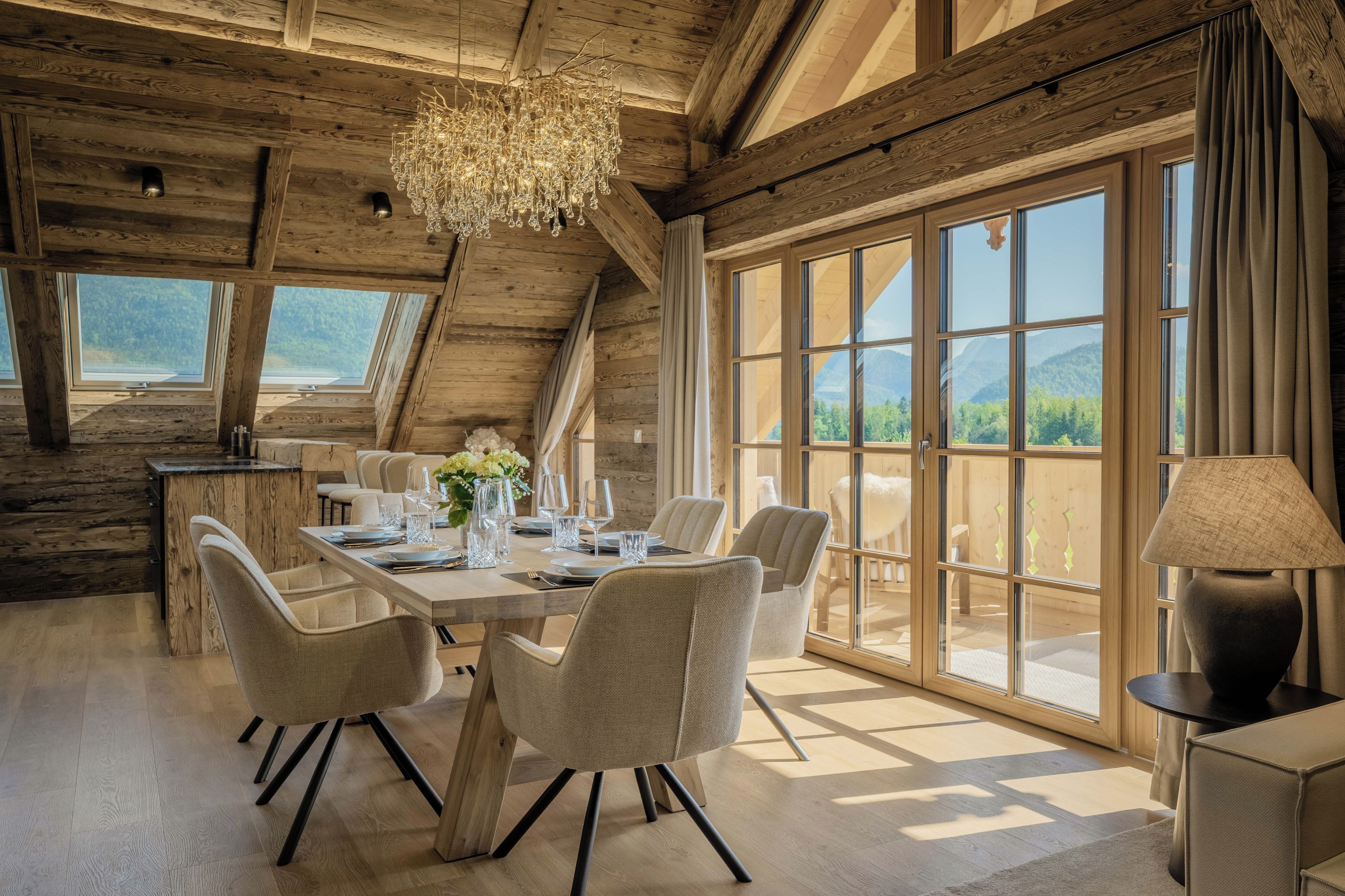 A dining area with a wooden table set for six, surrounded by chairs, with a large window offering a view of the outdoors.