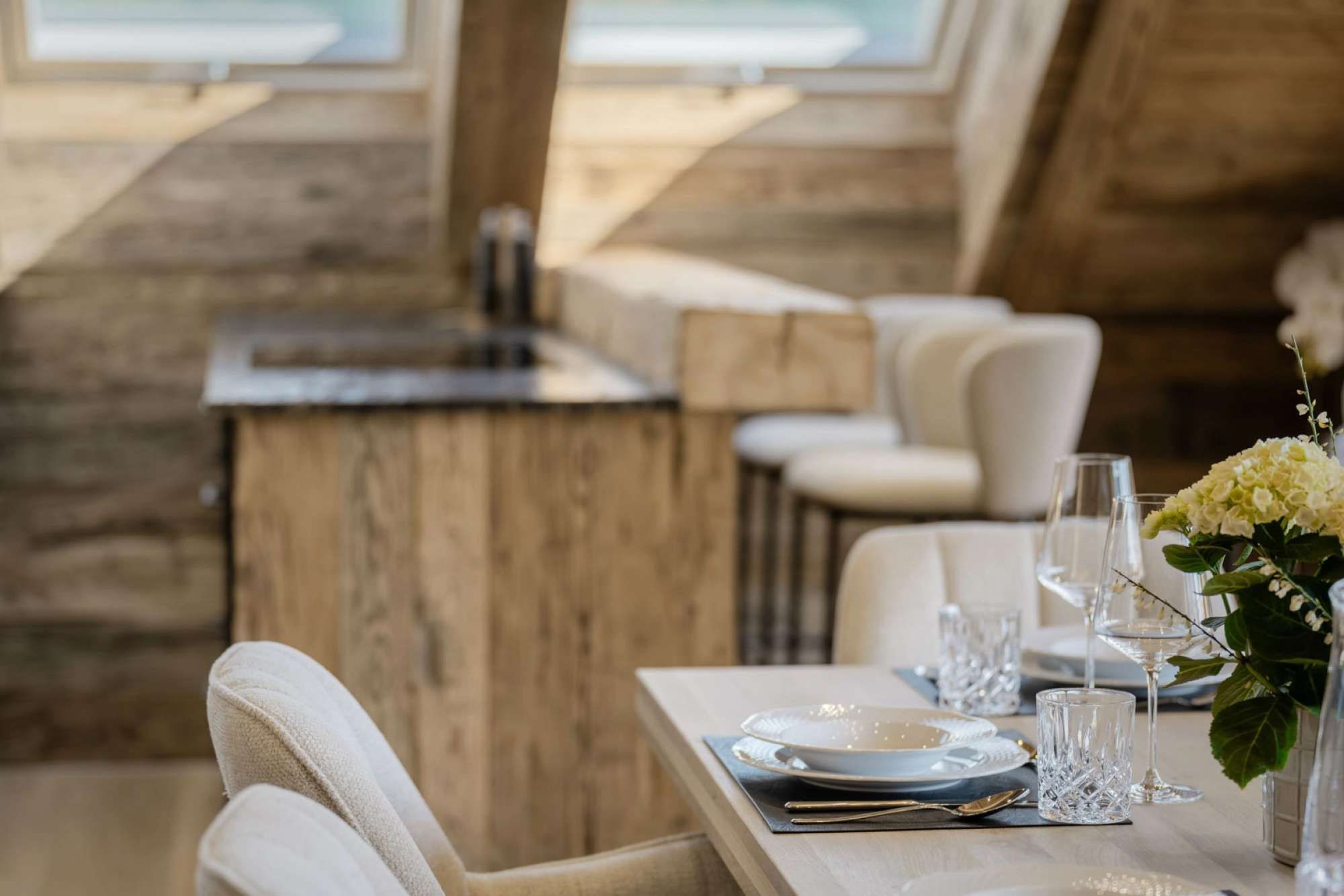 A dining area with a wooden table set for two, white chairs, and a vase with white flowers.