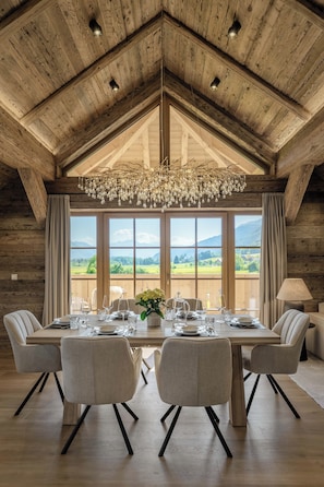 Ein Esszimmer mit großem Tisch, gedeckt für sechs Personen, Holzbalken an der Decke und Blick auf die Landschaft durch große Fenster.