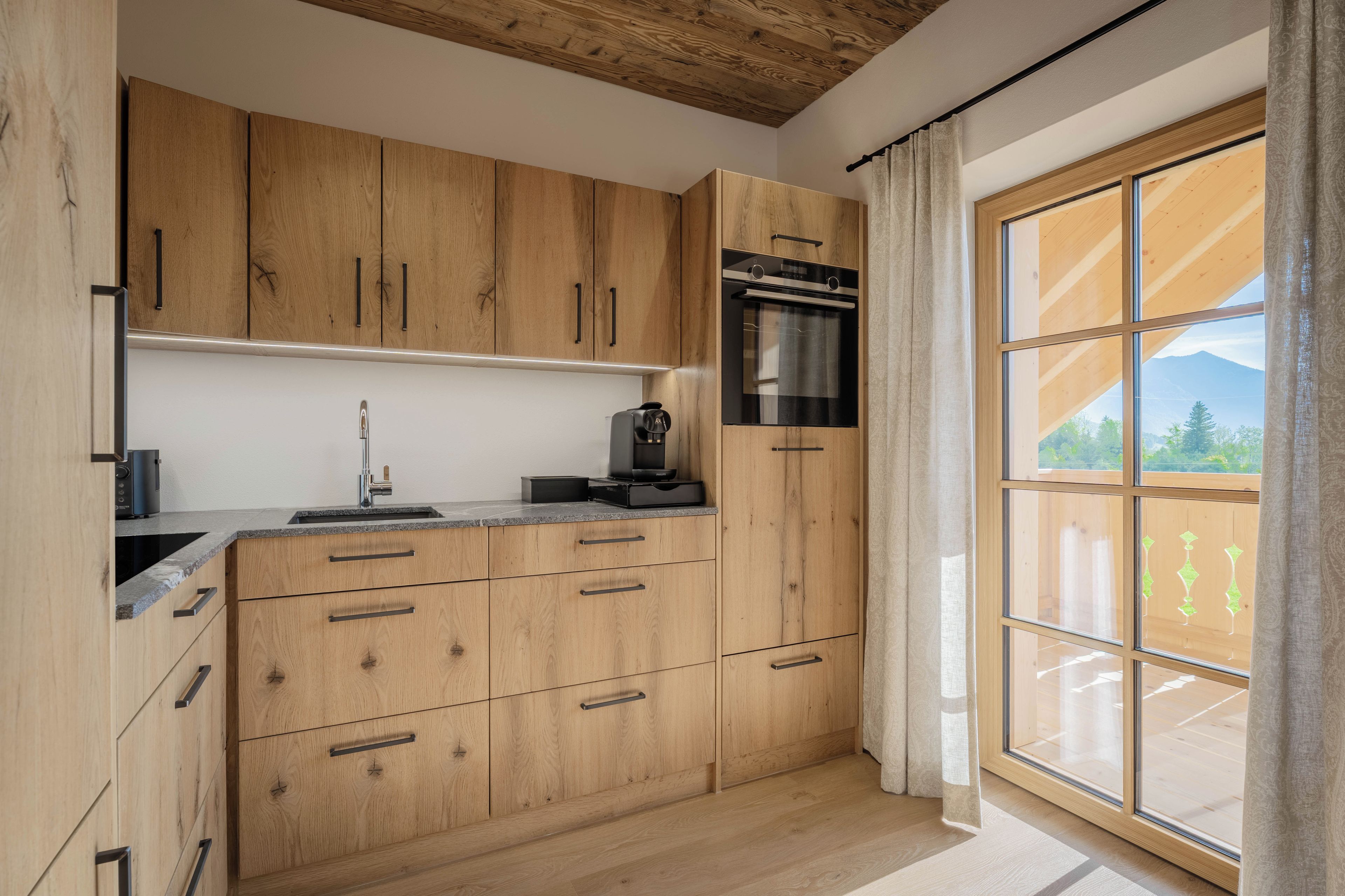 A kitchen with wooden cabinets, a stainless steel oven, a sink, and a window with a view of greenery and mountains.