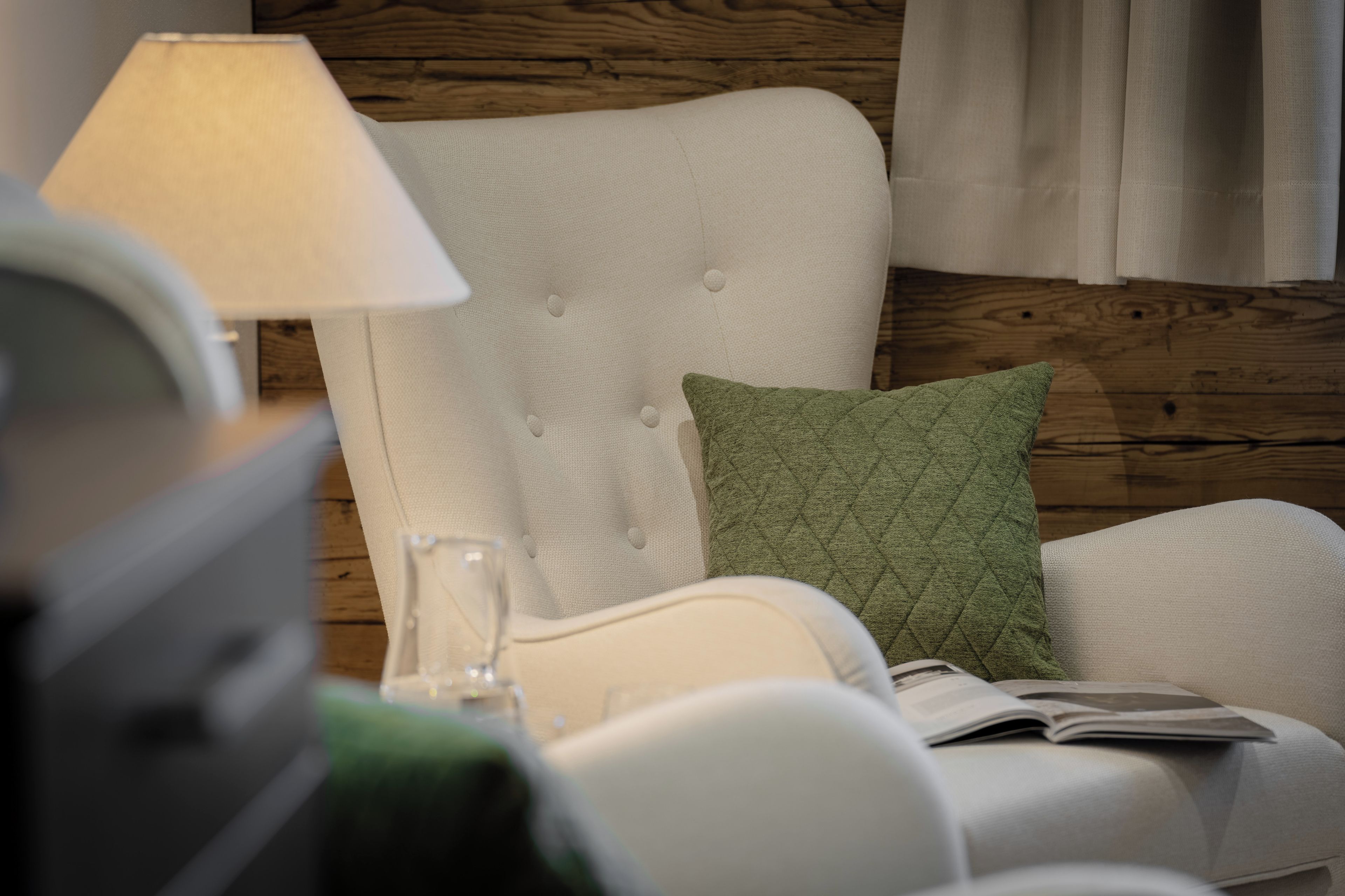 A room with a white upholstered chair, a green quilted pillow, a lamp, and a glass on the table.
