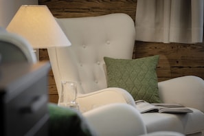 A room with a white upholstered chair, a green quilted pillow, a lamp, and a glass on the table. - Chalet Double Room for 2 Guests at Lake Wolfgang | spatzenNEST (Strobl)