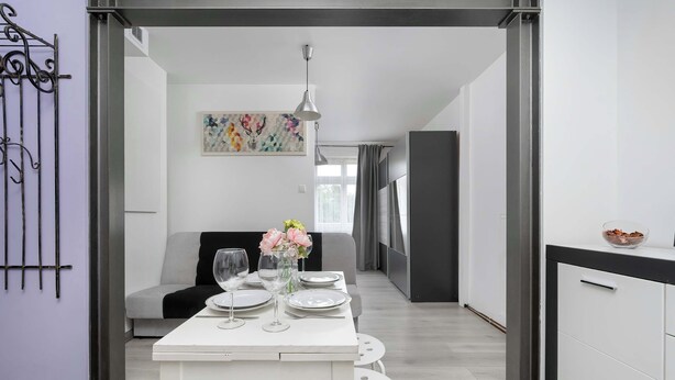 A modern dining area with a white table set for four, a grey sofa, and a decorative wall piece. - Lilac Studio with Bathtub | Close to the Park (Wrocław)