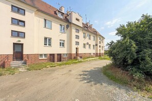 Un edificio a più piani con una base in mattoni e piani superiori color beige, un vialetto sterrato e un grande albero alla destra.