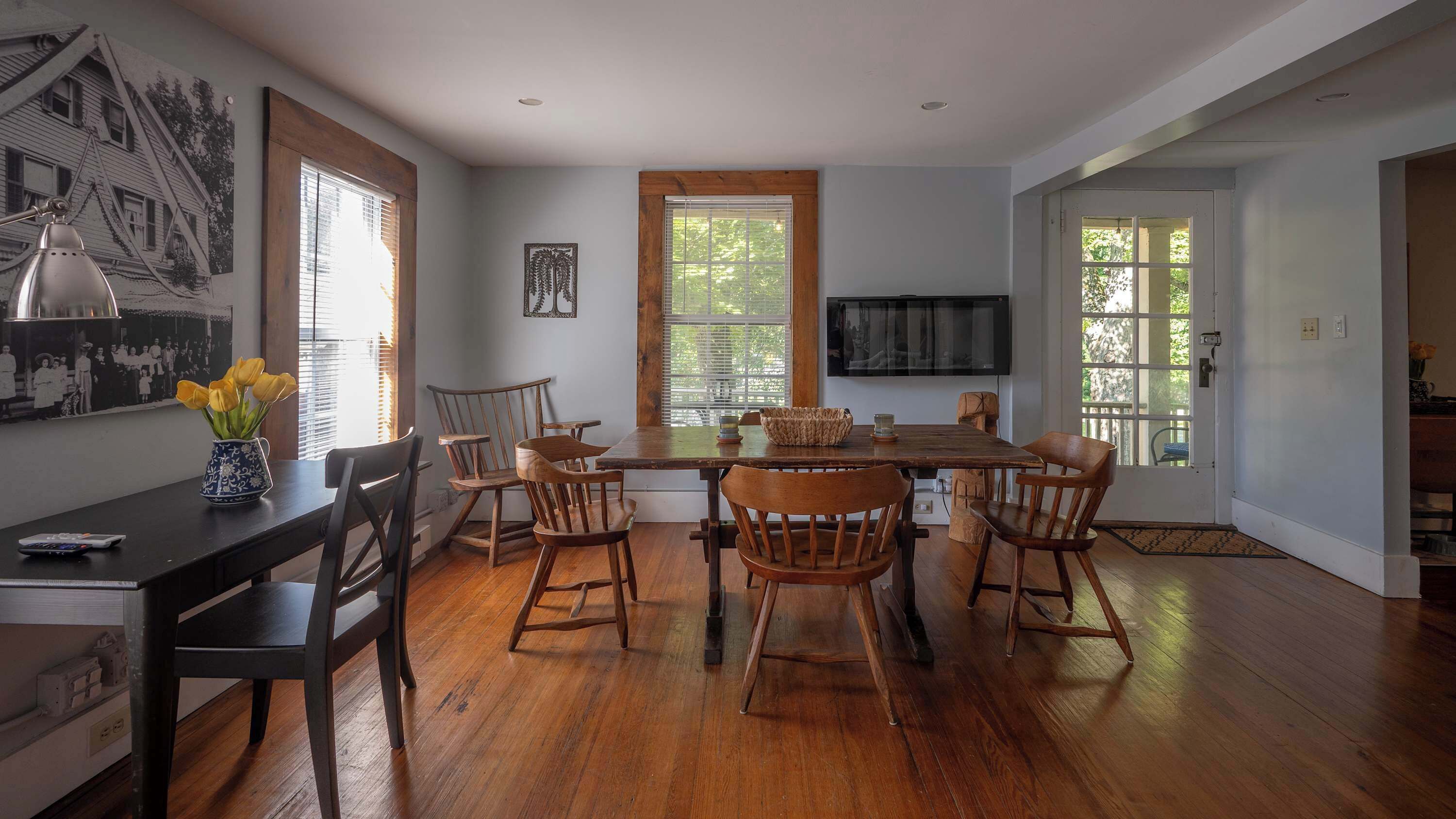 A dining area with a wooden table and chairs, a vase with yellow flowers, and a wall-mounted television.