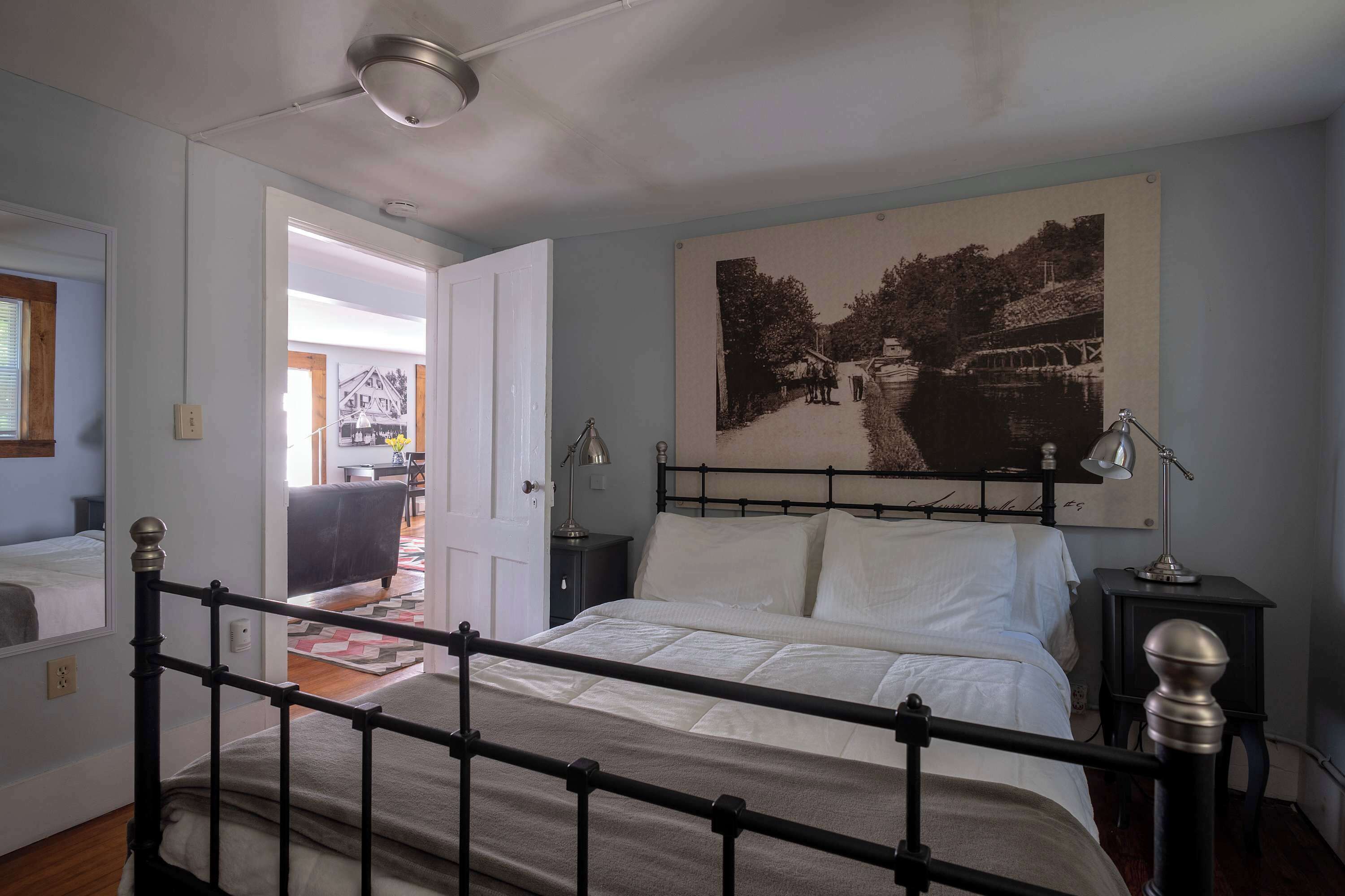A bedroom with a bed, bedside lamps, a nightstand, a painting on the wall, and a view of the outside.