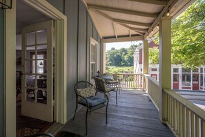 A covered porch with a table and chairs, a view of a green landscape, and a building with a red door.