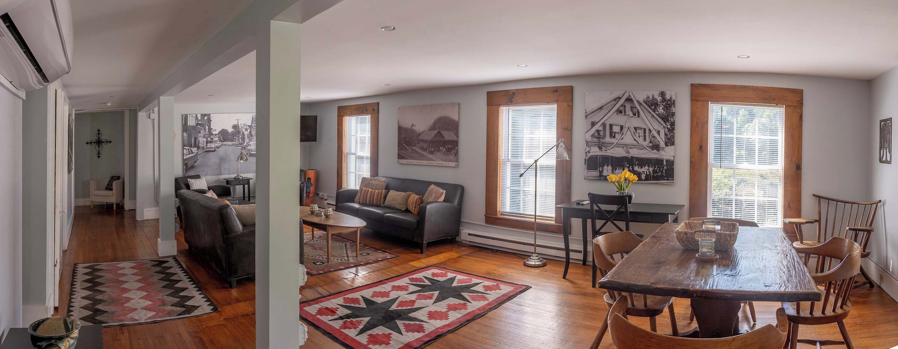A spacious living area with a sofa, a dining table, and a coffee table. There are large windows with blinds, a fireplace, and a wall-mounted TV.