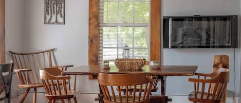 A dining area with a wooden table and chairs, a television mounted on the wall, and a window with blinds.