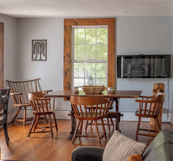 A dining area with a wooden table and chairs, a television mounted on the wall, and a window with blinds.
