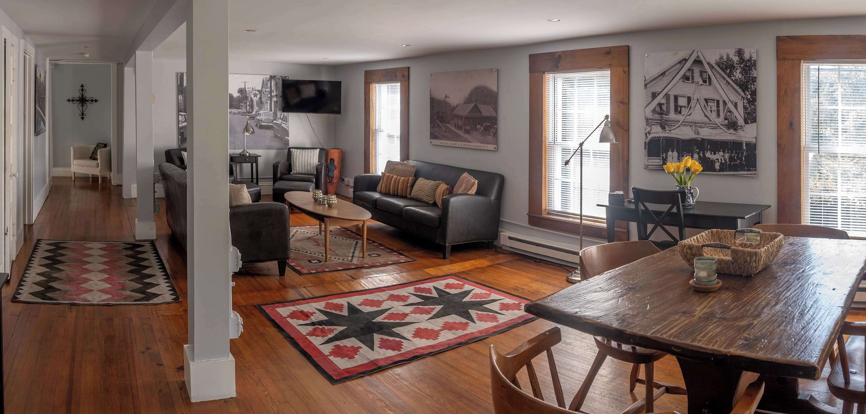 A spacious living area with a sofa, coffee table, and a dining table with chairs. There are framed pictures on the walls and a large window with blinds.