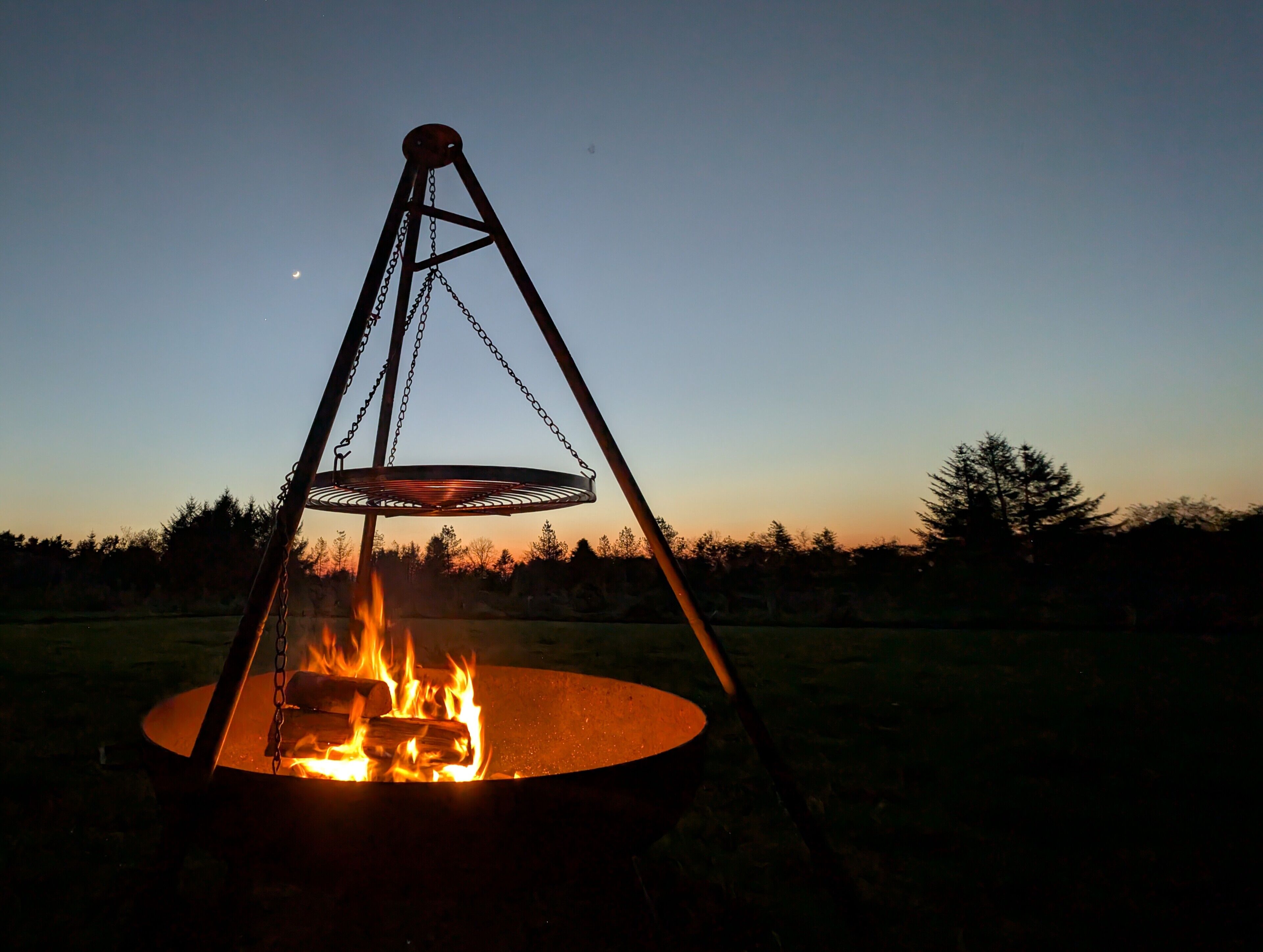 A fire pit with a metal frame and a circular fire ring, surrounded by a grassy field with trees in the background.