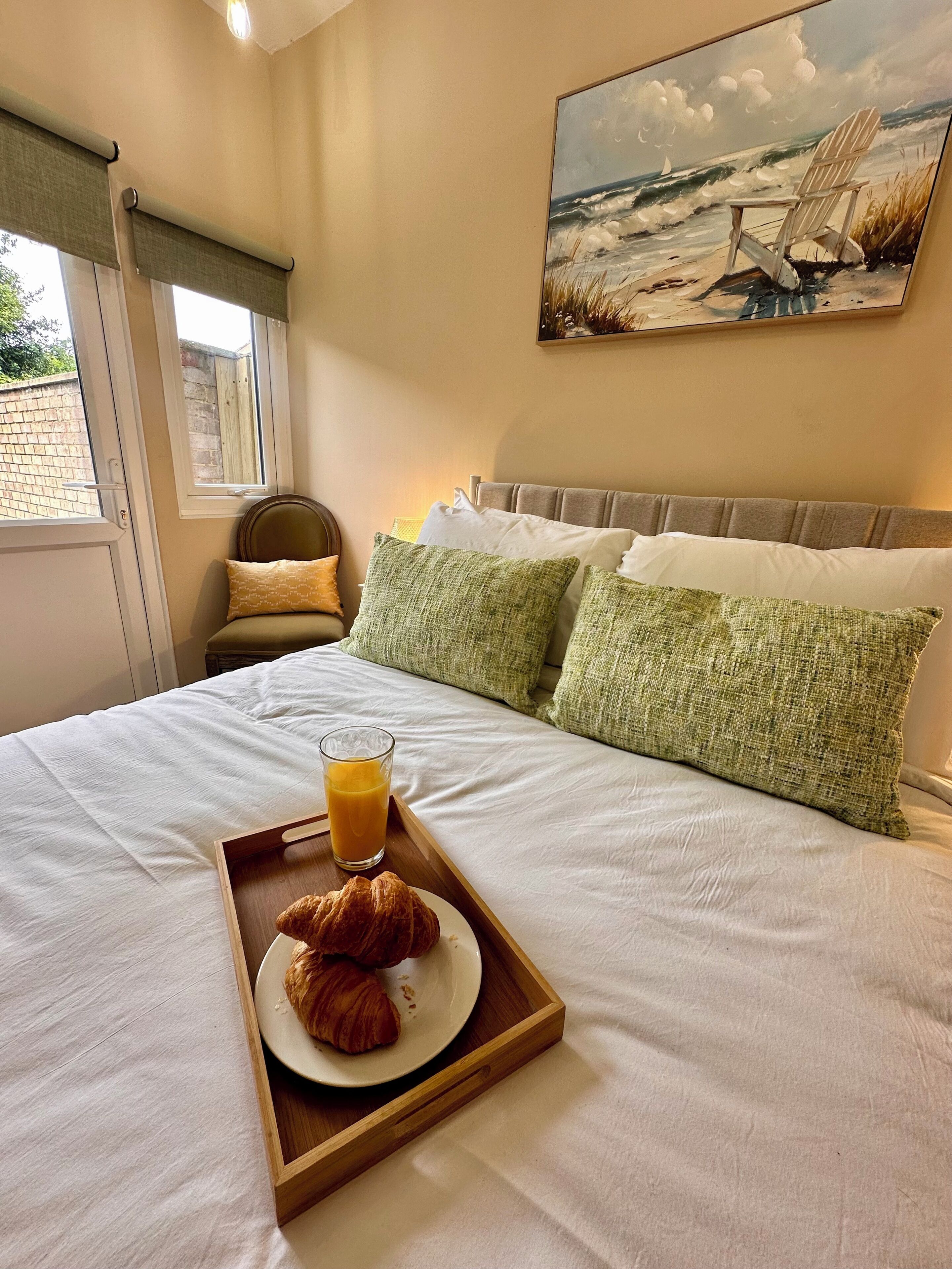 Un lit bien fait, avec un plateau contenant des croissants et du jus d’orange, un tableau représentant une scène de plage et une fenêtre avec des rideaux.