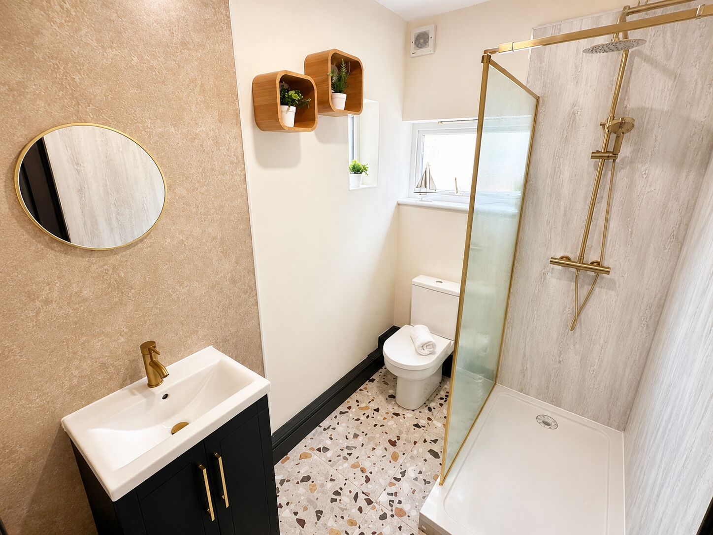 A bathroom with a white sink, a round mirror, a toilet, a shower with a glass door, and two wooden shelves with plants.